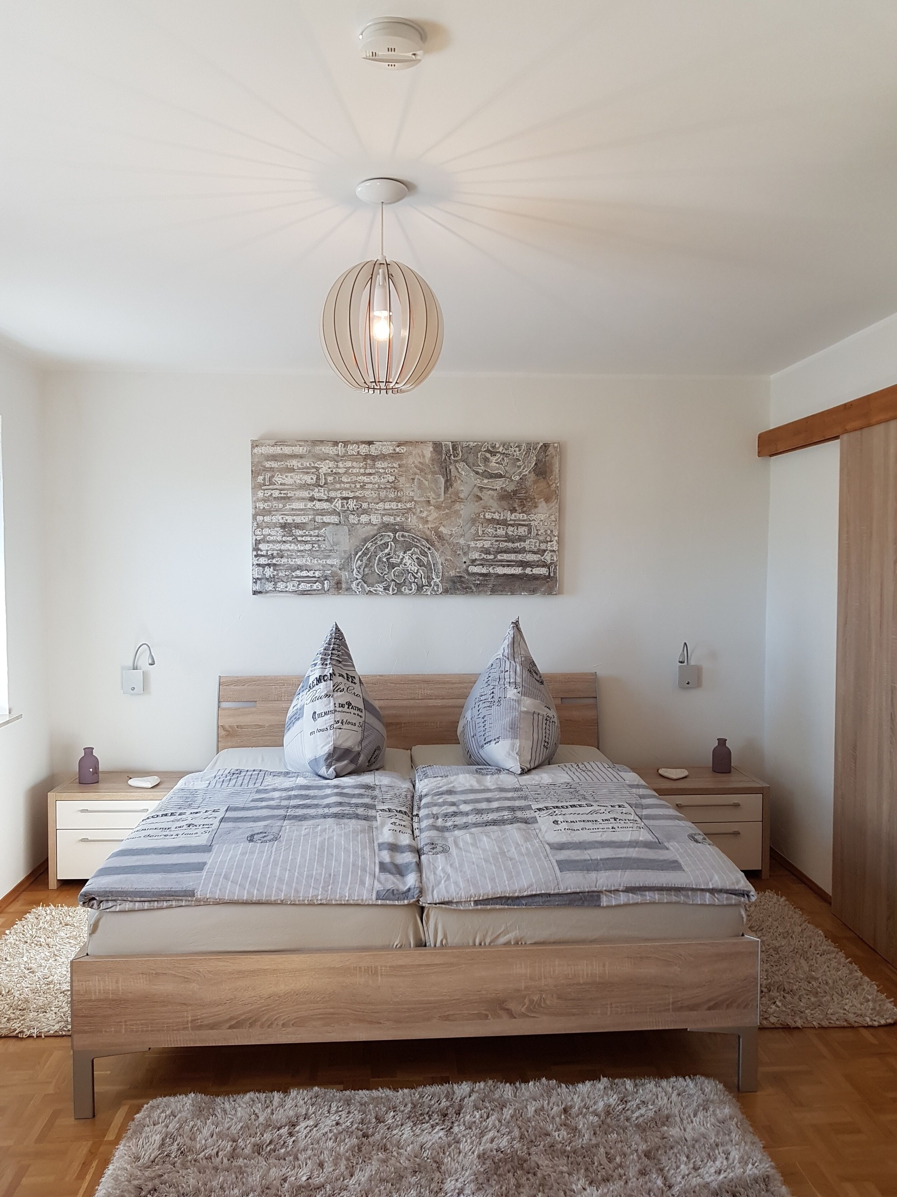 A bedroom with a wooden bed, two nightstands, a wall-mounted artwork, and a ceiling light fixture.