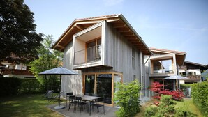 A modern two-story house with a patio area featuring a table and chairs, surrounded by a well-maintained lawn and a small garden with red-leaved plants.