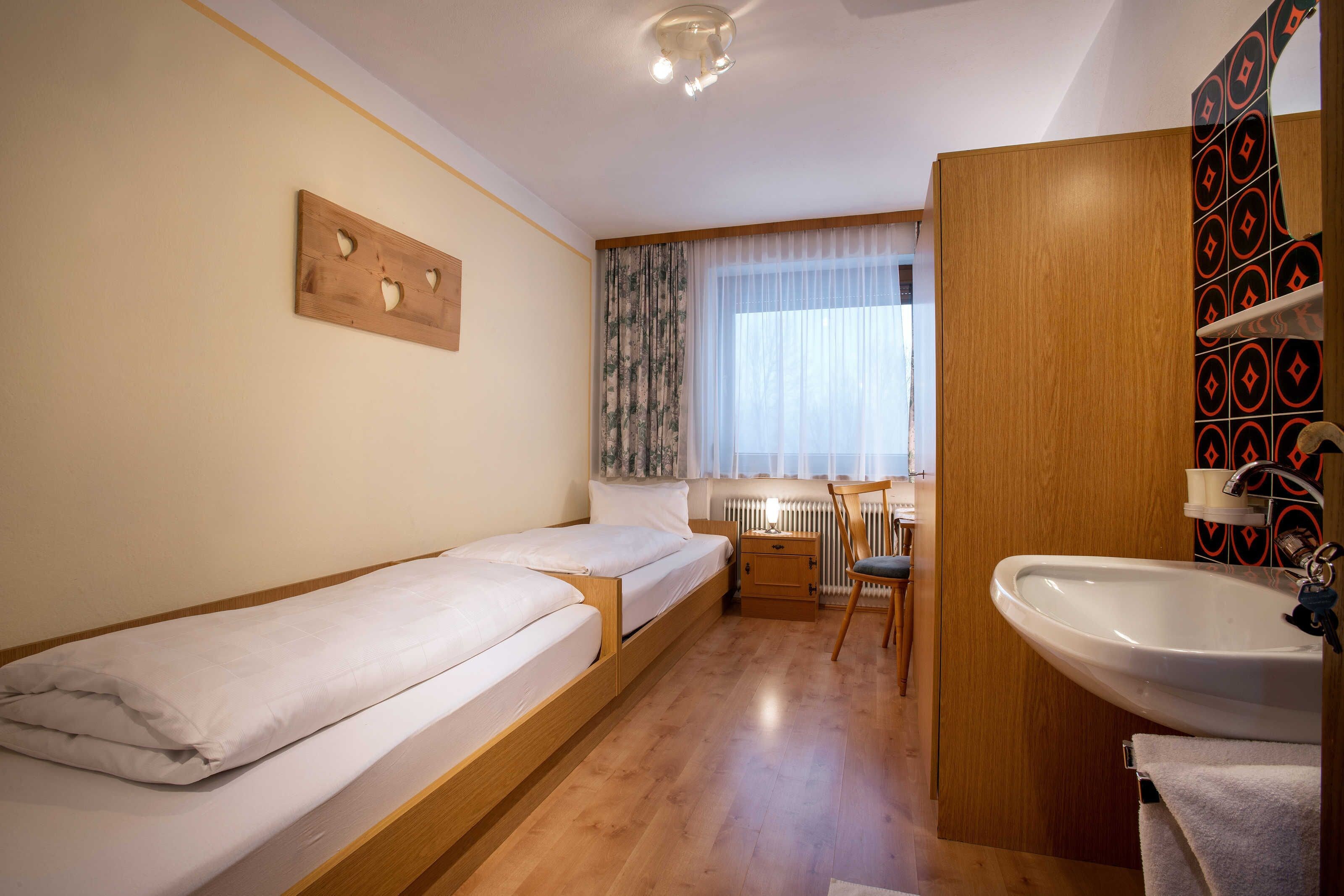 Una habitación pequeña y funcional con dos camas individuales, un escritorio de madera, una silla, un radiador y un baño con lavamanos y tendedero para toallas.