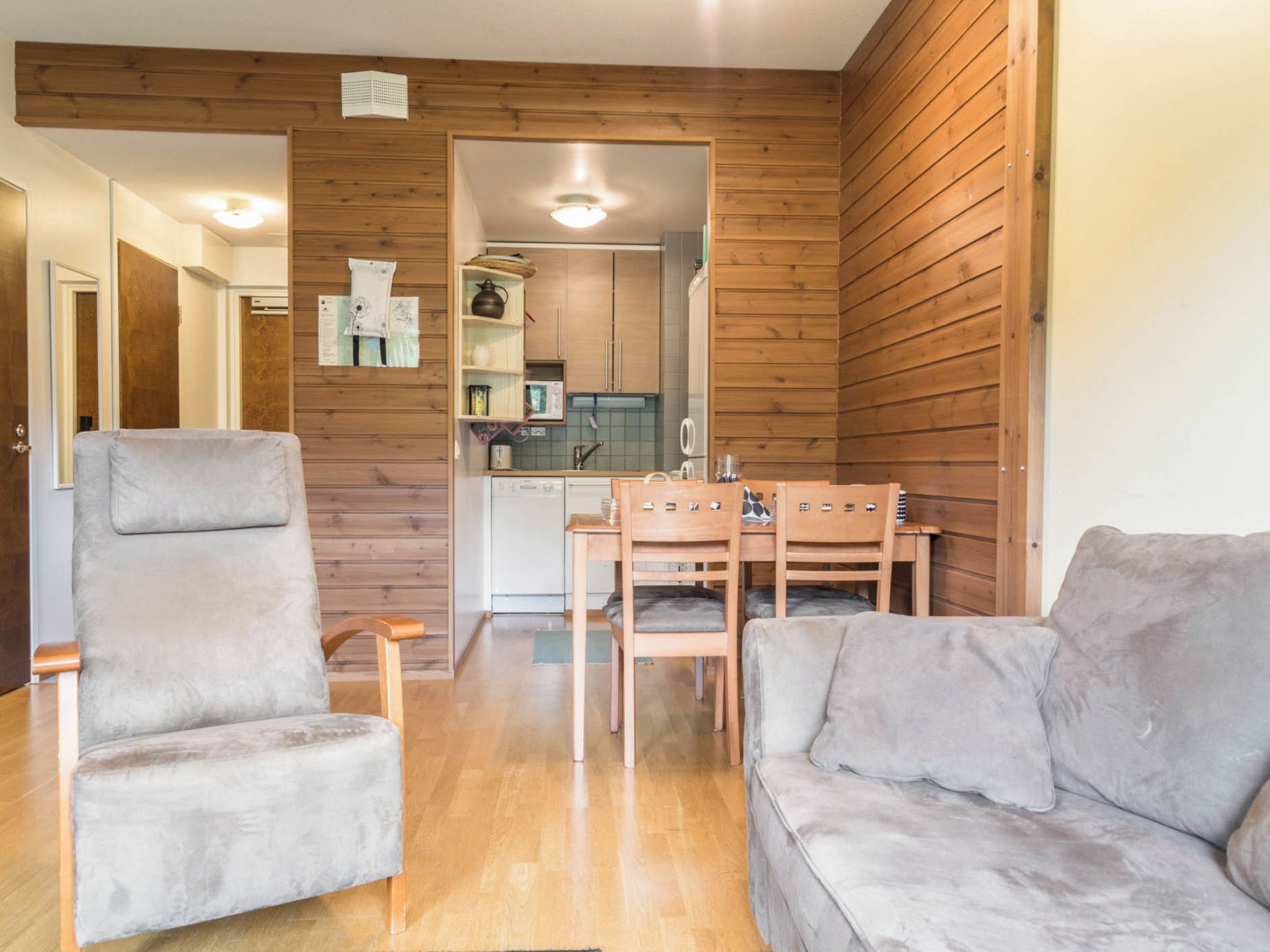 A room with a wooden wall, a grey sofa, a wooden dining table with chairs, and a kitchen area visible through an open door.