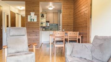 A room with a wooden wall, a grey sofa, a wooden dining table with chairs, and a kitchen area visible through an open door.