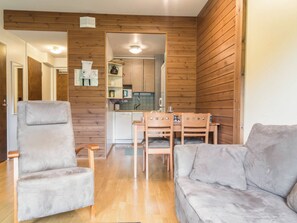 A room with a wooden wall, a grey sofa, a wooden dining table with chairs, and a kitchen area visible through an open door.