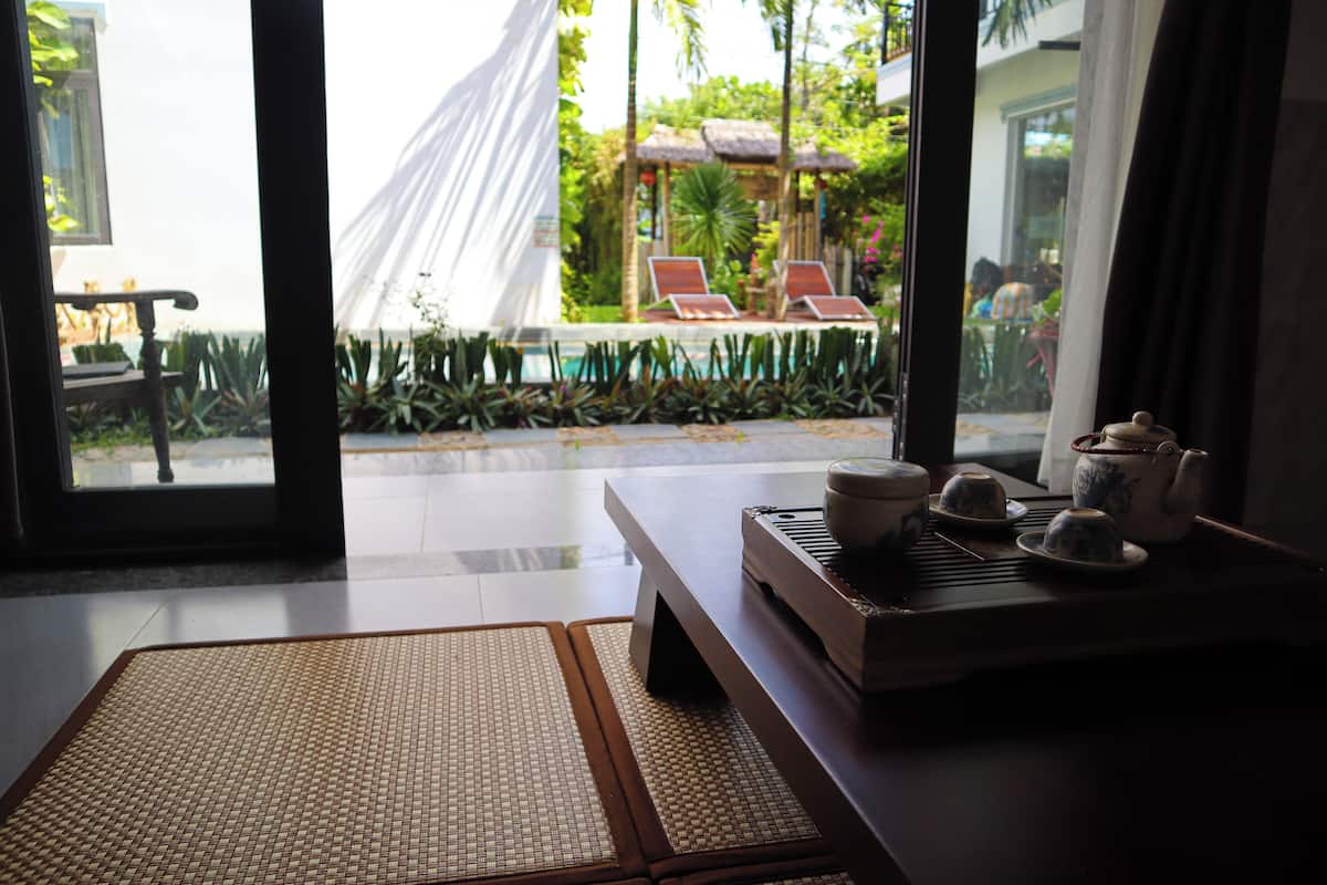 A room with a view of a pool area, a table with teapots, and a woven mat on the floor.