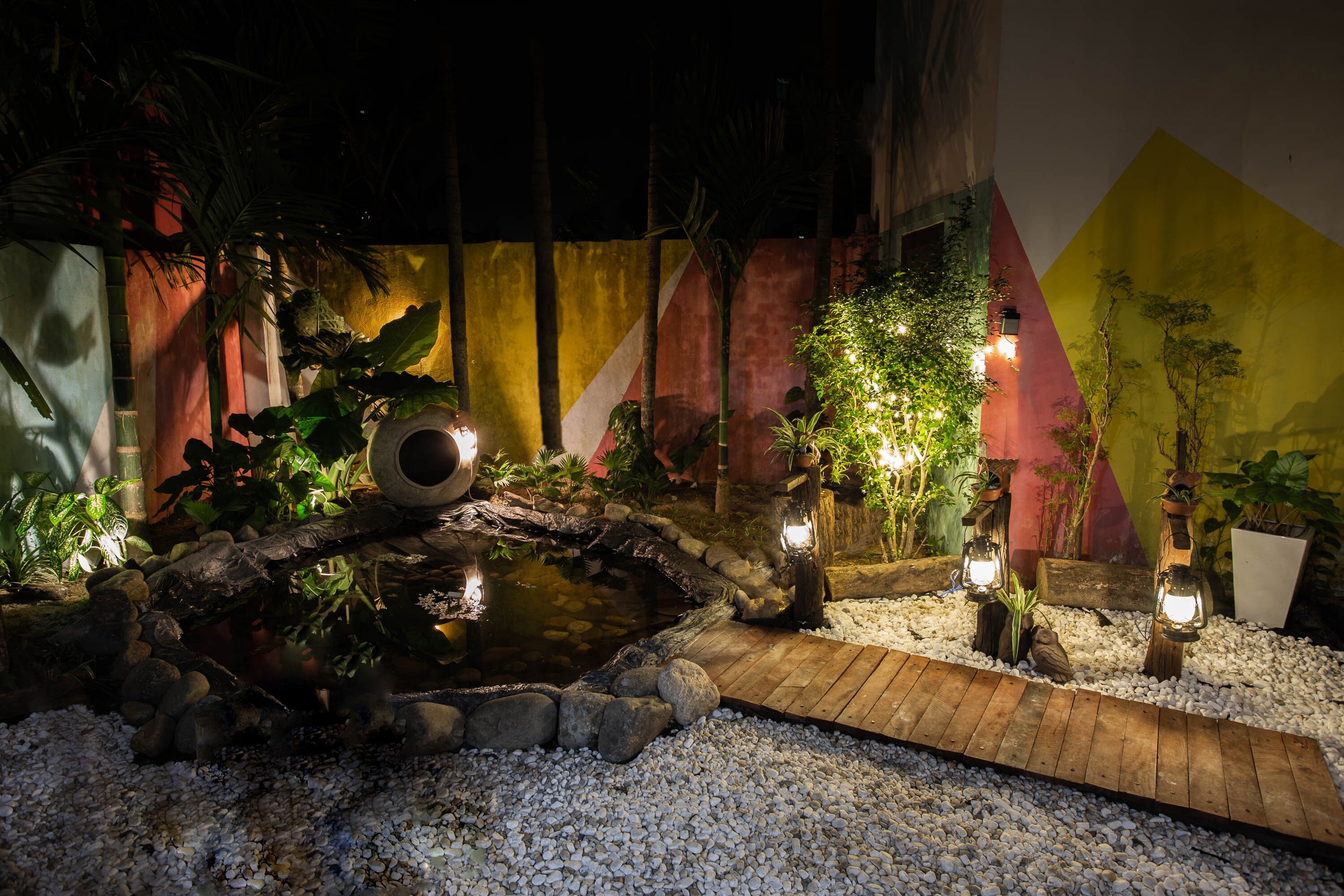 A nighttime garden with a small pond, wooden bridge, and various plants illuminated by lights.