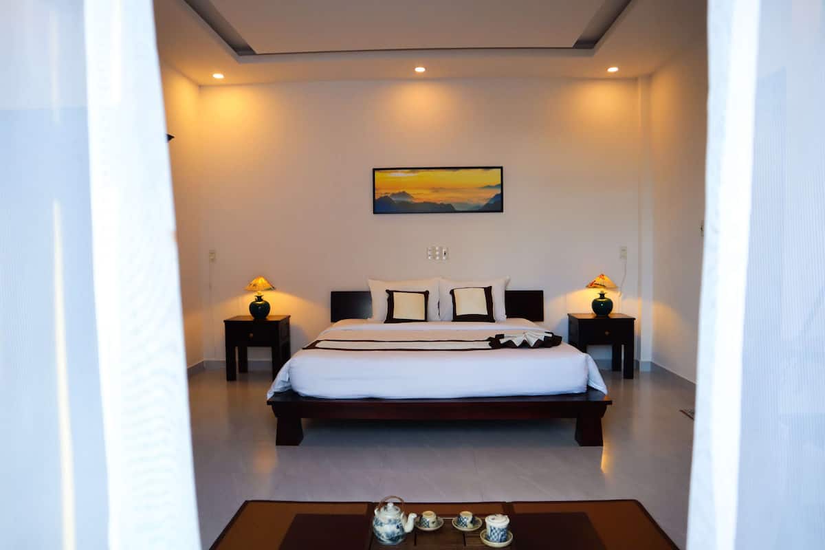 A bedroom with a large bed, two bedside tables with lamps, a painting on the wall, and a tea set on the floor.