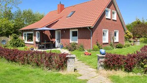 Ein zweistöckiges Backsteinhhaus mit rotem Ziegeldach, einer überdachten Terrasse mit Gartenmöbeln und einem gepflegten Garten mit roten Sträuchern.
