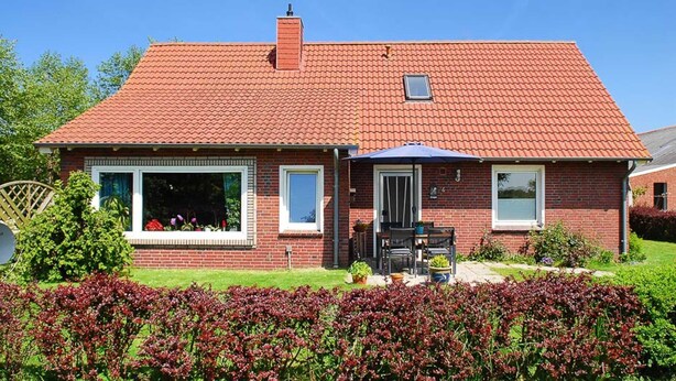 Ein zweistöckiges Backsteinhäuschen mit rotem Ziegeldach, einer Veranda mit Tisch und Stühlen sowie einem kleinen Garten mit Sträuchern mit roten Blättern.