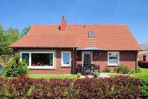 Une maison à deux étages, construite en briques, avec un toit recouvert de tuiles rouges, un porche à l’avant doté d’une table et de chaises, et un petit jardin agrémenté d’arbustes aux feuilles rouges.