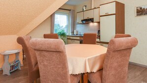 Ein Essbereich mit einem runden Tisch und sechs Stühlen, ein kleiner Beistelltisch mit Büchern und eine Küche im Hintergrund.
