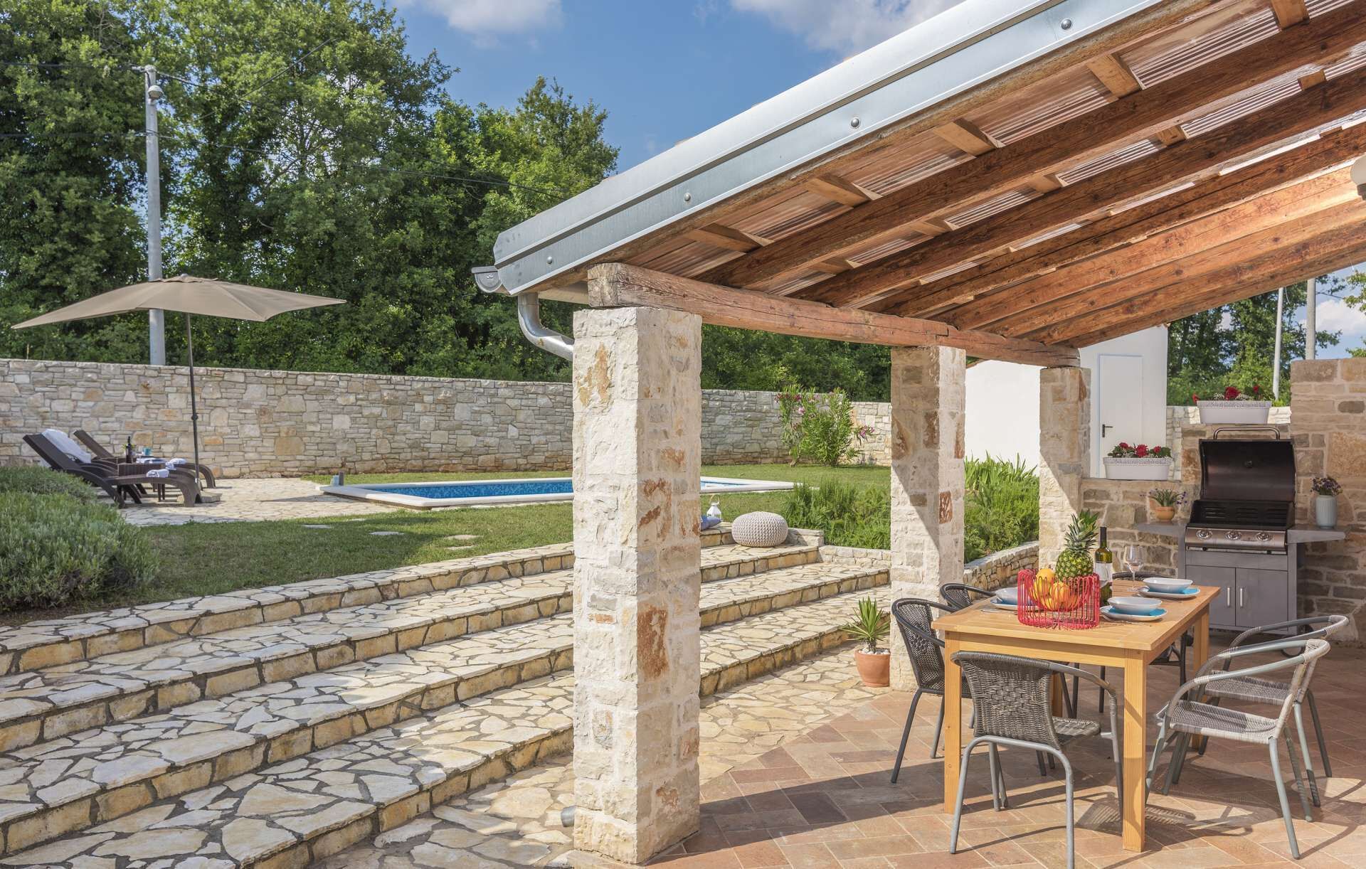 A covered outdoor patio with stone steps, a pool, lounge chairs, and a barbecue area.