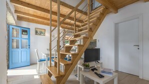 A wooden staircase leading to a second floor, a dining area with white chairs and a blue door, and a small table with a plant and remote controls.