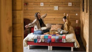 Een knus houten chaletinterieur met een bed, voorzien van een kleurrijke quilt, en twee personen die erop zitten.