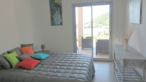 Un dormitorio con cama, almohadas coloridas, un cuadro, un balcón con vistas y una mesita con un jarrón.