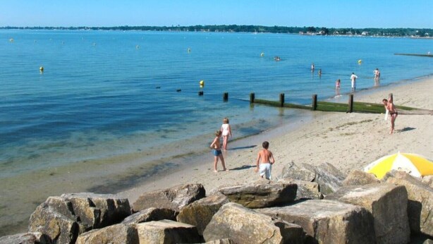 Une plage avec une eau d’un bleu limpide, des gens qui se promènent et de gros rochers au premier plan.