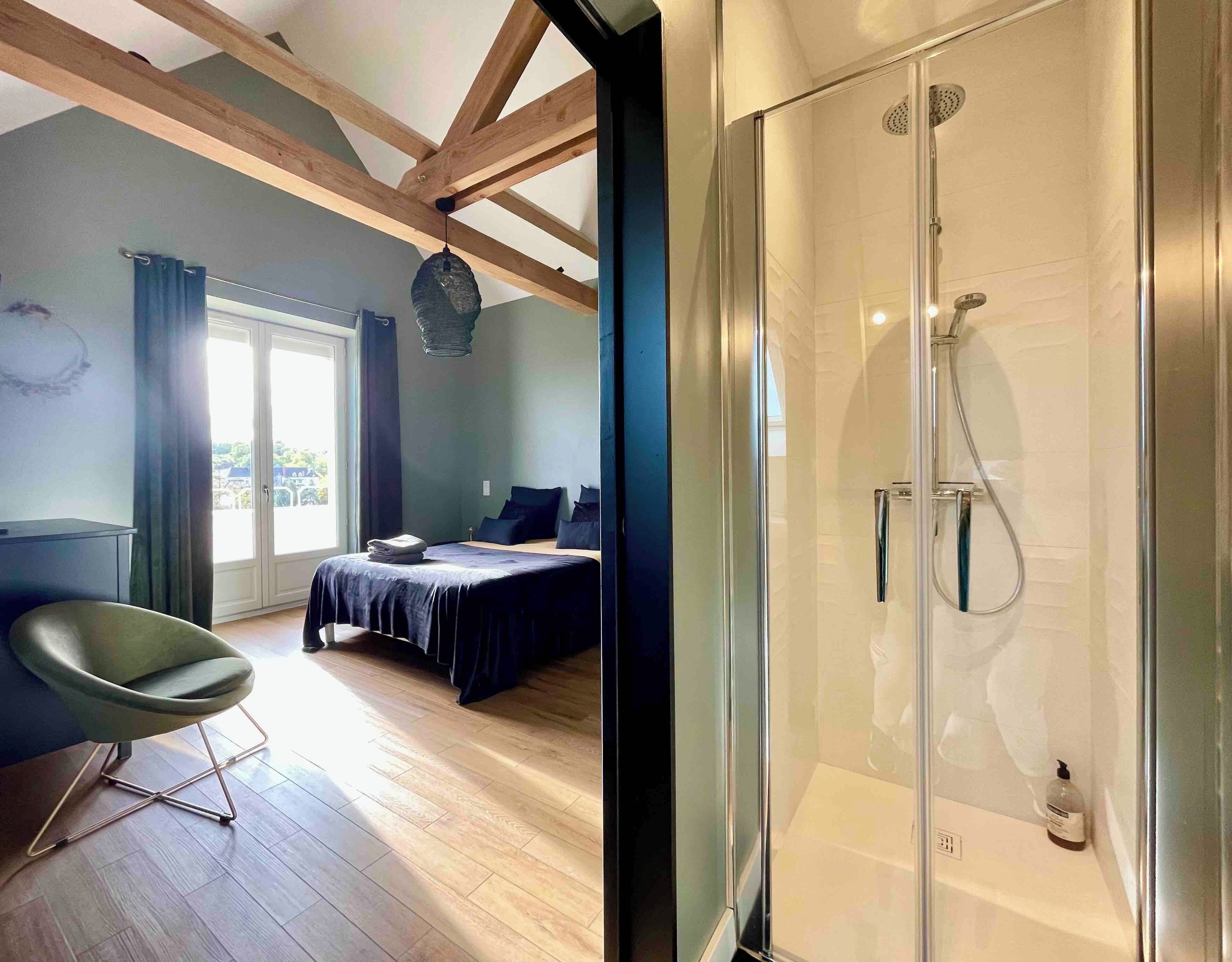 A modern bedroom with a glass shower, a bed with dark bedding, a green chair, and a view through a large window.