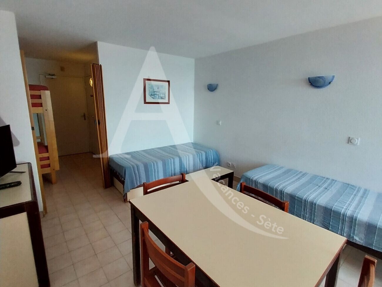 A small, compact room with a single bed, a desk, and a chair. There is a framed picture on the wall and a small shelf with decorative items.
