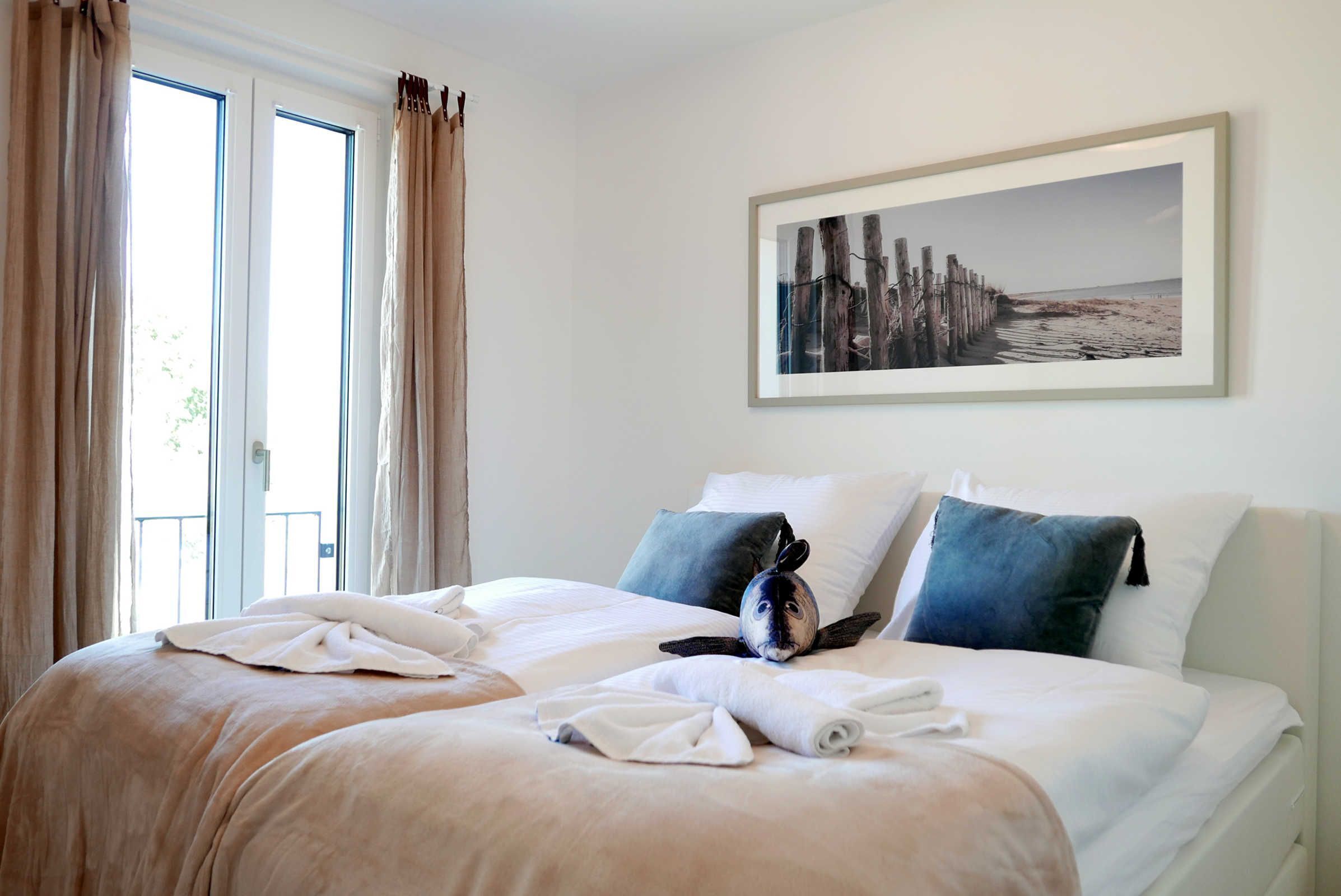 A hotel room with two beds, a large window with curtains, a framed picture on the wall, and a towel on the bed.