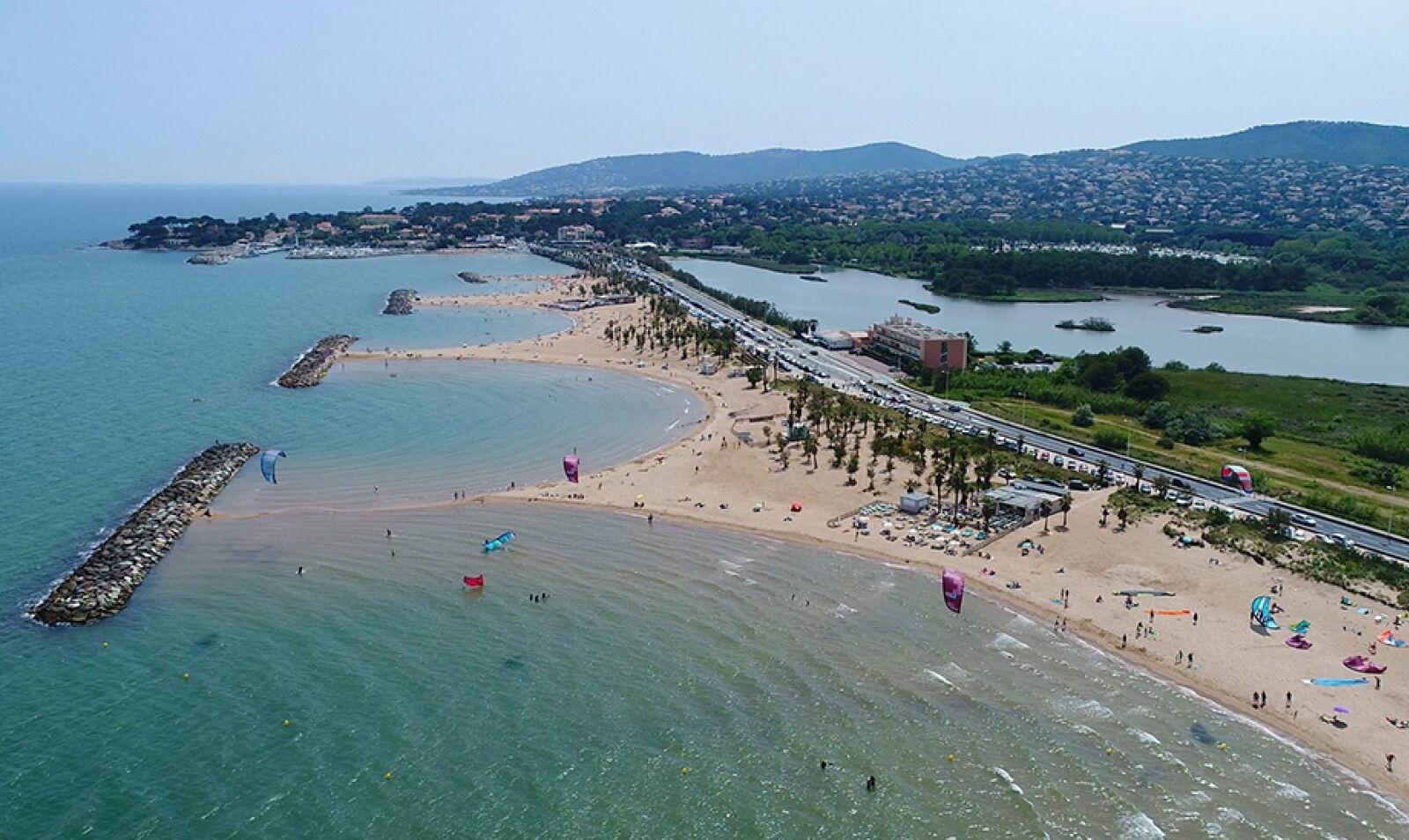Vue aérienne d’une zone côtière avec une plage, une promenade et un paysage urbain en arrière-plan.