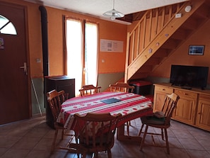 A dining area with a table set for four, wooden chairs, a television on a wooden cabinet, and a staircase leading to an upper level.