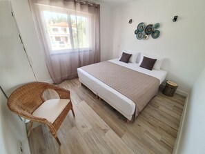 Quarto com cama, cadeira de vime, janela com cortinas e um objeto decorativo na parede.