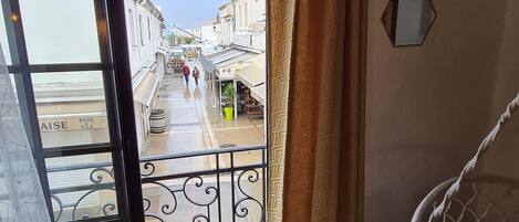 Una habitación con vista a una calle estrecha, un balcón con baranda de hierro forjado y una cama con almohadones estampados.