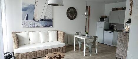 Un espacio habitable moderno con un sofá blanco, una mesa de comedor y sillas, una zona de cocina con electrodomésticos, y un reloj grande en la pared.