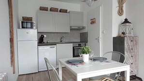 Cozinha compacta com armários brancos, micro-ondas, lava-louças e forno pequeno. Há uma mesa de jantar com pratos e um vaso de planta.