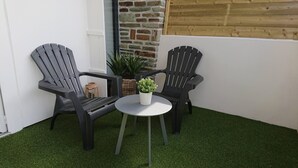 Uma pequena área de estar ao ar livre com duas cadeiras pretas e uma mesa branca, cercada por grama sintética.