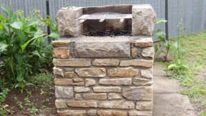 Un barbecue extérieur en pierre, doté d’une grille en métal, entouré de verdure et d’une clôture.