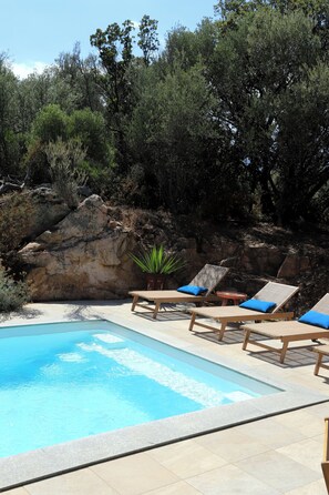 Piscina con sillas de descanso y una pequeña mesa, rodeada de un paisaje rocoso y árboles.