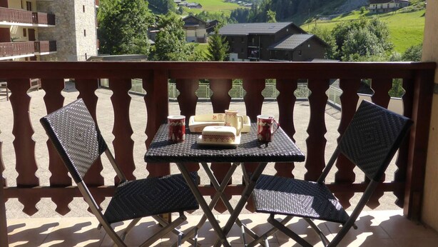 Ein Balkon mit zwei Stühlen und einem Tisch, mit Blick auf eine gebirgige Landschaft und Häuser.