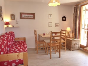 A cozy living room with a red patterned sofa, wooden dining table with chairs, a small side table with a lamp, and a wicker basket.