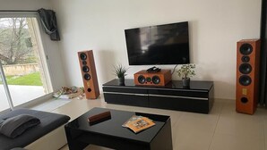 Ein modernes Wohnzimmer mit einer schwarzen Couch, einem Couchtisch mit einer Zeitschrift, einem Fernseher an der Wand und zwei großen Lautsprechern.