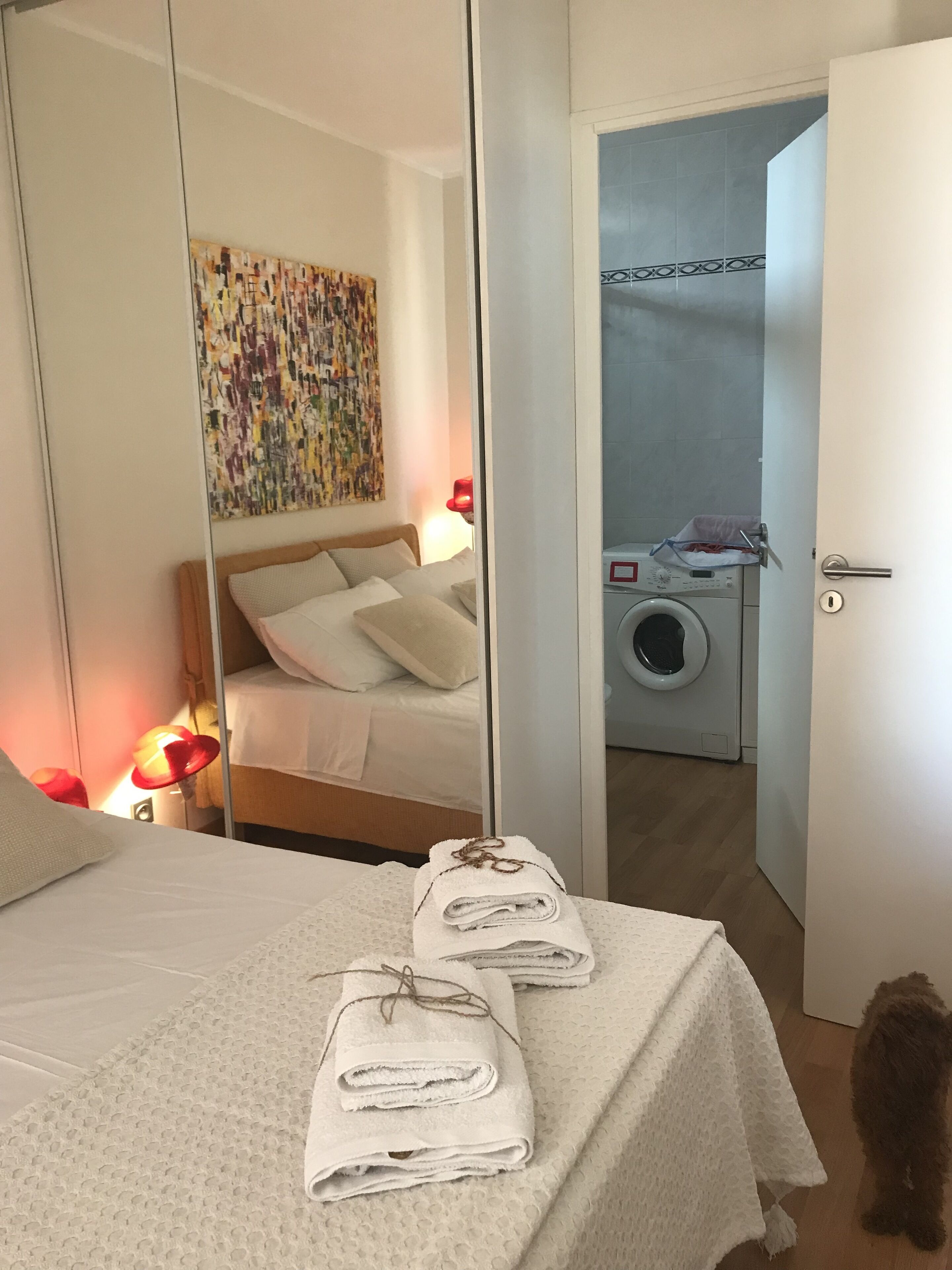 Quarto de hotel com cama, espelho, obra de arte na parede, máquina de lavar e porta-toalhas.