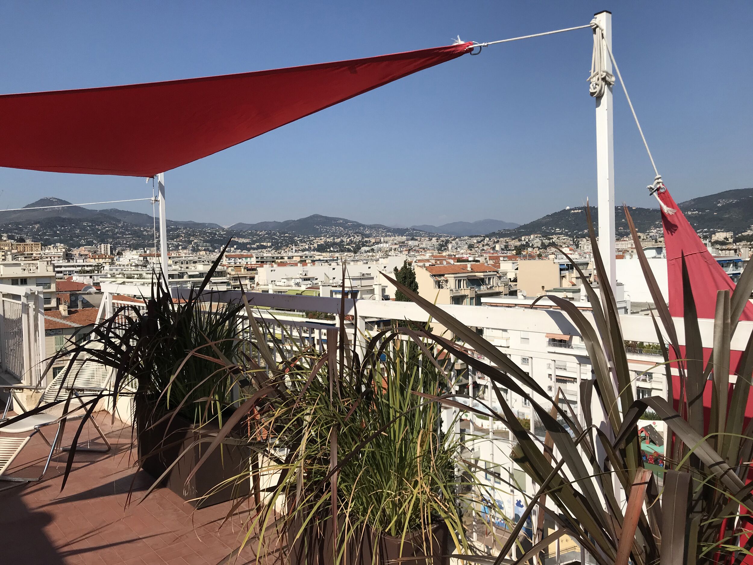 A terrace with a red awning, potted plants, and a view of a cityscape with buildings and hills in the background.