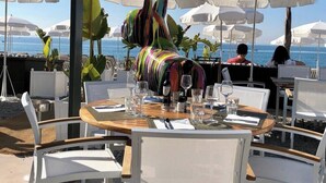 Un espace de restauration en plein air avec des chaises et des tables blanches, des parasols blancs et une vue sur l’océan.