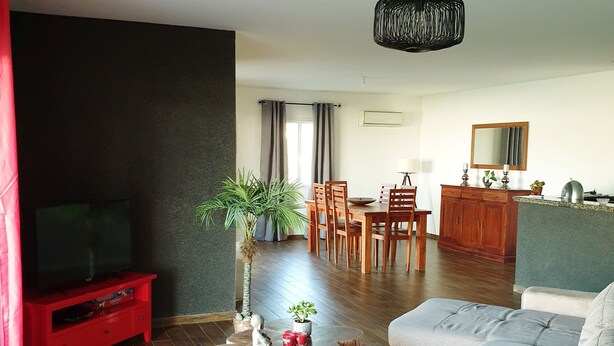 Ein modernes Wohnzimmer mit einem grauen Sofa, einer roten Unterhaltungsanlage, einem Holztisch und einem Essbereich im Hintergrund.