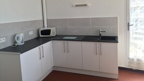 Uma cozinha compacta com armários brancos, bancada preta, micro-ondas, chaleira e janela com vista para prédios.