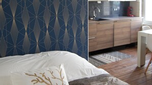 Ein modernes Hotelzimmer mit Wand im geometrischen Muster, einem Bett mit weißem und goldenem Zierkissen, Holzboden, einer Küchenzeile mit weißen Schränken und einem weißen Stuhl.