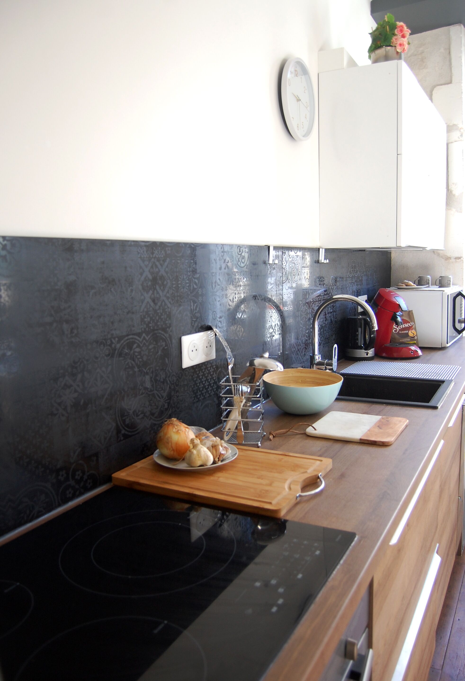 Une cuisine moderne avec une plaque de cuisson noire, un plan de travail en bois, un évier et une horloge au mur.