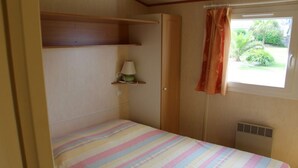 A small, single-bed room with a striped bedspread, a bedside lamp, a shelf with a lamp, and a window with curtains. - CHALET 12 ILLIADE PANORAMIC SEA VIEW, (Plomodiern)