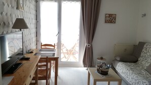 A living room with a TV, wooden furniture, a table with a basket, a sofa with a patterned cover, and a view of a balcony. - very nice studio DINARD / LA RICHARDAIS 5 minutes from the beach (LA RICHARDAIS)