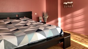A bedroom with a geometric-patterned bedspread, a dark headboard, a bedside table with a lamp, and a wall-mounted decorative piece. - The lodge "L'Allée du Val" (Lalaye)
