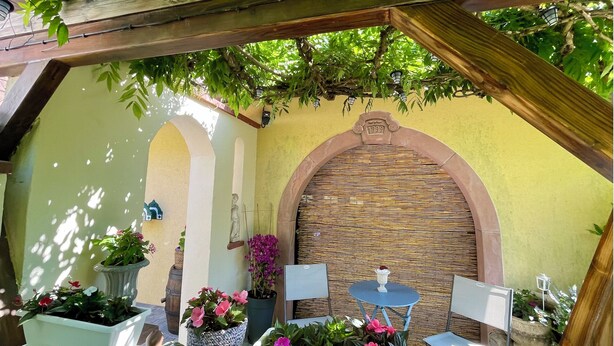 A cozy outdoor seating area with wooden tables and chairs, surrounded by potted plants and a decorative wall. - The lodge "L'Allée du Val" (Lalaye)