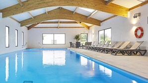 Une piscine intérieure dotée d’un plafond en bois, de nombreuses fenêtres et de chaises longues disposées le long des bords.
