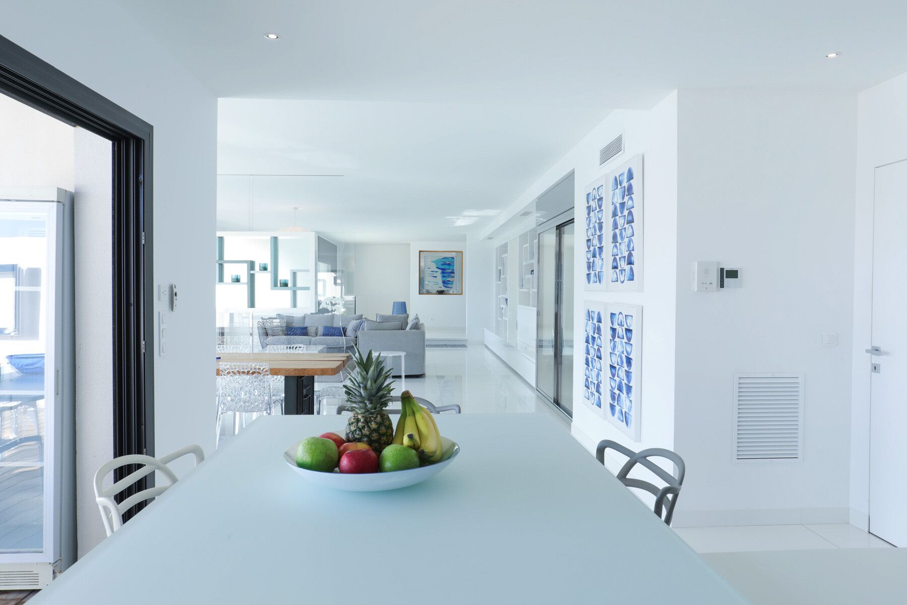 A modern kitchen with a long dining table, a fruit bowl, and a view into the living area.