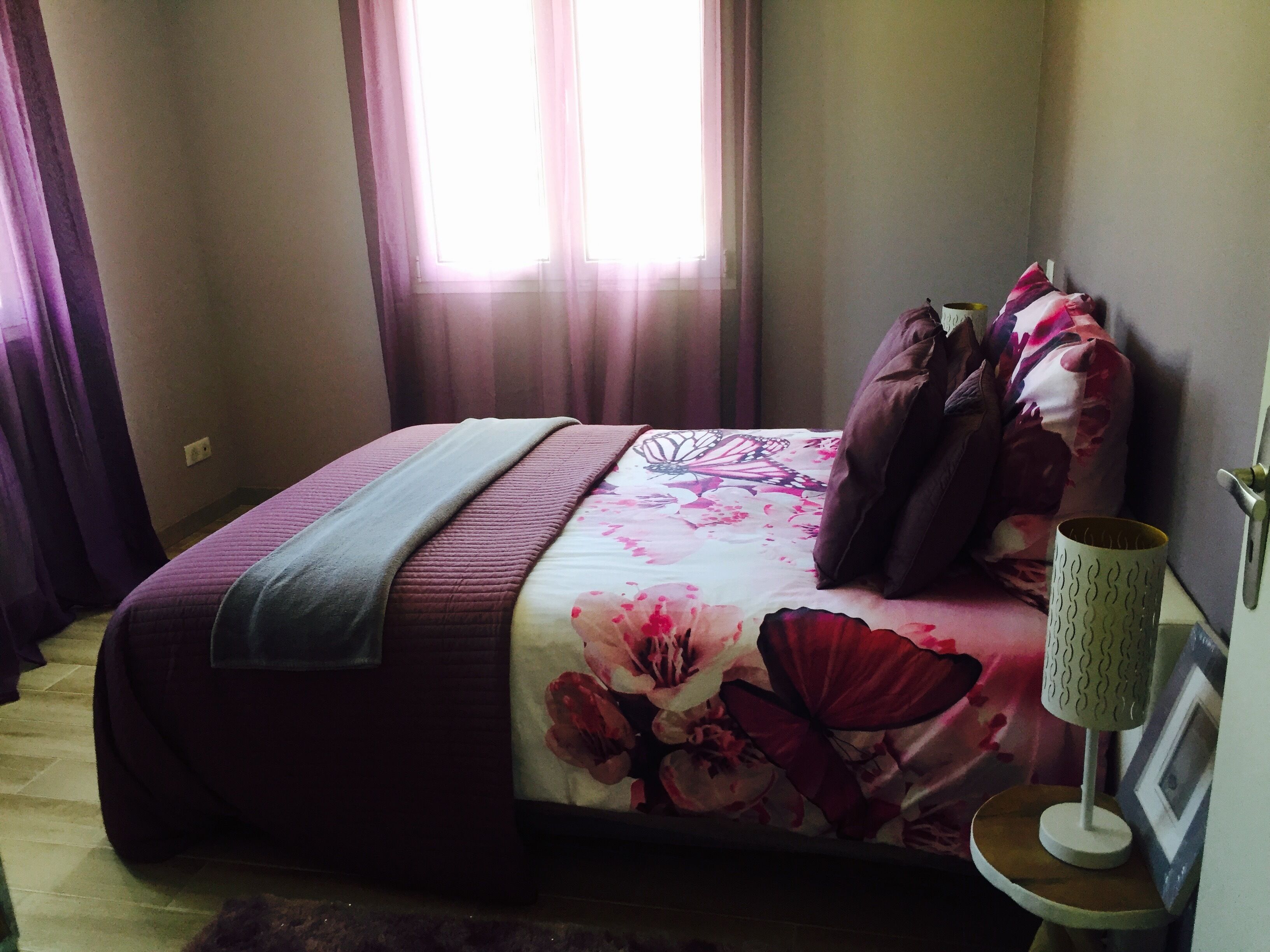 Un dormitorio con cama, cortinas, una lámpara de noche y un cuadro en la pared.