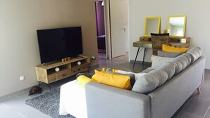 Un salon moderne avec un canapé gris, des coussins jaunes, un téléviseur à écran plat posé sur un meuble en bois, une petite table d’appoint et un miroir avec un cadre jaune.
