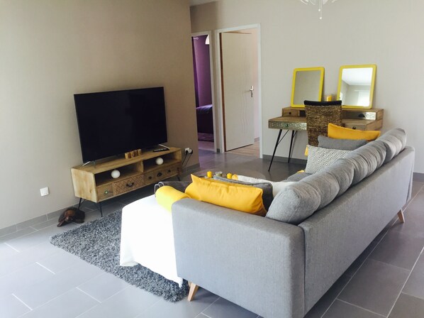 A modern living room with a grey sofa, yellow cushions, a flat-screen TV on a wooden stand, a small side table, and a mirror with yellow frames. - Brand new holiday villa  (Bompas)