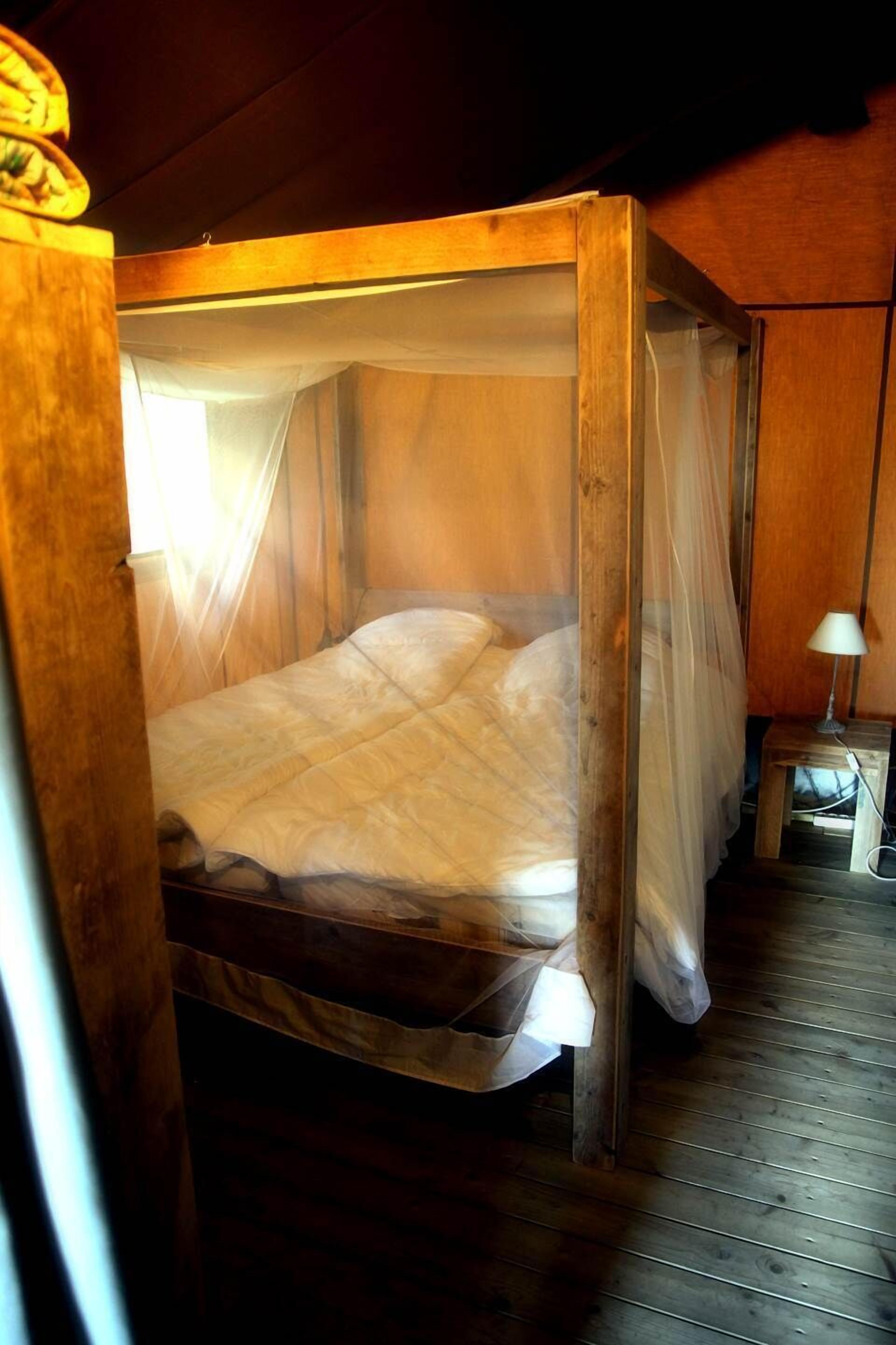 A wooden four-poster bed with a canopy, a bedside table with a lamp, and a wooden floor.